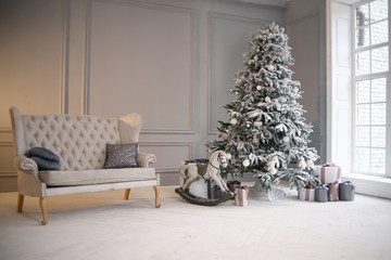 New Year's decor in a photo studio in gray and white colors. A snow-white tree, gifts and lanterns, against the background of a white wall. Horses, sofa and snow. Location for a photo shoot. Christmas
