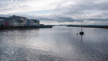 One sail boat on a calm Galway bay with the famous colorful houses & sunrise, ocean & cloudy sky in the distance. Taken early morning.
