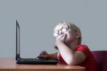 Pensive blond boy with glasses sits at a laptop. Internet and preschooler