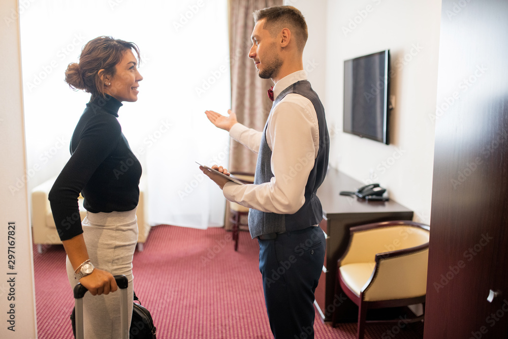 Poster young porter in uniform showing room to one of guests with baggage