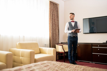 Contemporary young porter using touchpad while standing in one of hotel rooms