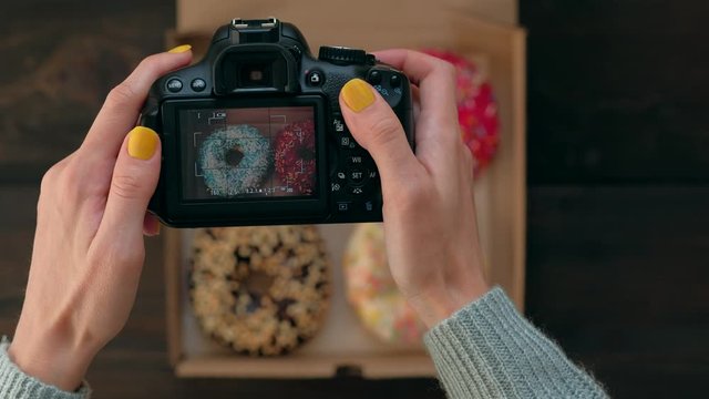 Top view - Woman's hands take pictures with photo dslr camera of delicious fresh donuts in box on wooden table. 4k. Concept of food photo blogging.