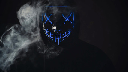Man with angry and scary lighting neon glow mask in hood on black background with smoke. Halloween...