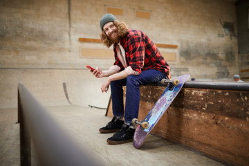 Full length portrait of contemporary bearded man looking at camera and smiling while using smartphone siting on ramp in skating park, copy space