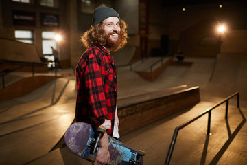 Waist up portrait of contemporary bearded skater looking at camera and smiling while posing in urban skating park lit by dim light, copy space
