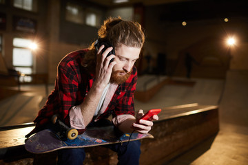 Portrait of contemporary bearded man listening to music via smartphone while sitting on ramp in dim skating park, copy space