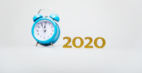 New Year is coming. A blue alarm clock with a black and white clock face, placed on a white background near the golden numbers of 2020.