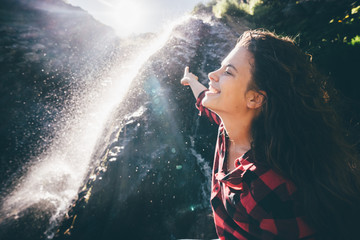 Young happy girl enjoing the waterfall. Woman standing in front of waterfall with rased hands and smile. Concept of ecotourism travel