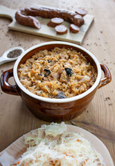 Bigos in stoneware dish, next to bigos ingredients: sausage, sauerkraut and cumin
