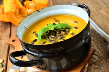 Enamel pot of cream pumpkin, carrot soup with fresh basil, pumpkin seed and laddle on rustic wooden table.