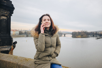 Fototapeta na wymiar Smiling tourist woman in Prague speaking on smartphone. Young woman tourist stands on the Charles Bridge in Prague in the Czech Republic uses phone