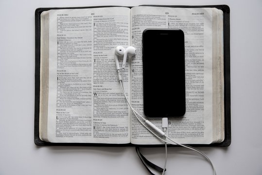 Overhead shot of a smartphone and headphones on an opened bible on a white surface