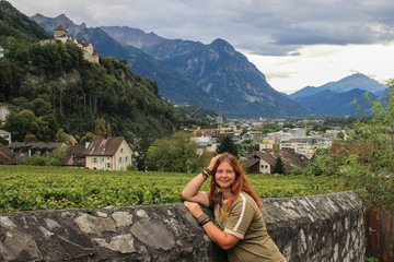 Naklejka na ściany i meble Caucasian young red-haired hippie girl tourist on the background of the panorama of Vaduz, Alps mountains and Vaduz Castle, Liechtenstein - the official residence of the prince.