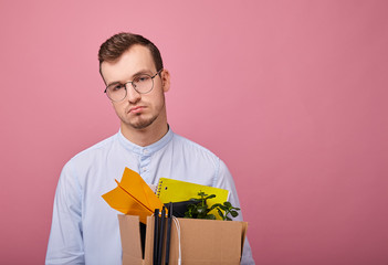 A pretty guy in a sky blue shirt and computer glasses stands on a pink background with a cardboard box with different things in his hands. Dismissal. Fired.