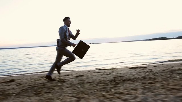 Happy businessman holding in hand briefcase and running on the beach at sunset.