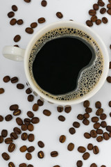 Vertical photo of black aromatic coffee with froth in a white mug among coffee beans in defocus on a white background. View from above