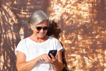 Fototapeta premium Cheerful senior lady using mobile phone to send a message. Smiling and writing. Sunset light. Background of rusty sheet metal