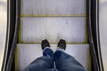 Male feet in jeans and black sneakers standing on escalator POV