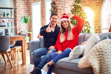 Young couple wearing santa claus hat sitting on the sofa around christmas tree at home smiling making frame with hands and fingers with happy face. Creativity and photography concept.
