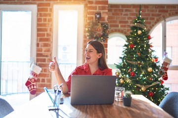 Beautiful woman sitting at the table working with laptop at home around christmas tree Looking proud, smiling doing thumbs up gesture to the side