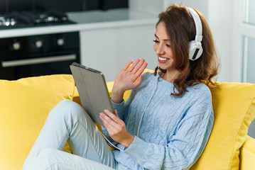Charming cheerful girl with headphones sitting on the couch, holding laptop or tablet pc and waving hands to her internet friend. Young woman communicates online with her friends abroad.