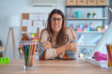 Young beautiful teacher woman wearing sweater and glasses sitting on desk at kindergarten shaking and freezing for winter cold with sad and shock expression on face
