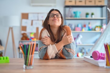 Young beautiful teacher woman wearing sweater and glasses sitting on desk at kindergarten Hugging...