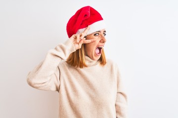 Beautiful redhead woman wearing christmas hat over isolated background Doing peace symbol with fingers over face, smiling cheerful showing victory