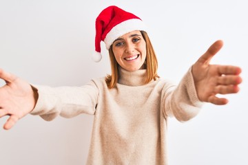 Beautiful redhead woman wearing christmas hat over isolated background looking at the camera smiling with open arms for hug. Cheerful expression embracing happiness.