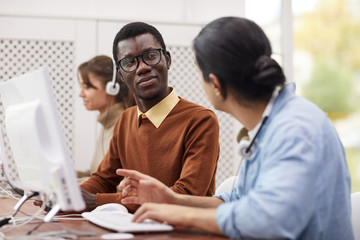 Portrait of African-American student wearing glasses talking to classmate while using computers in college library, copy space