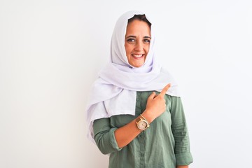 Young beautiful Arab woman wearing traditional Muslim hijab over isolated background cheerful with a smile of face pointing with hand and finger up to the side with happy and natural expression