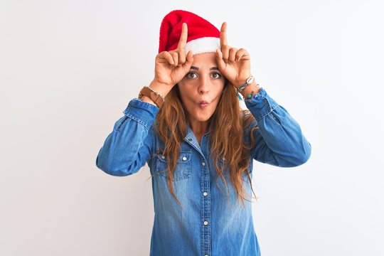 Young beautiful redhead woman wearing christmas hat over isolated background doing funny gesture with finger over head as bull horns