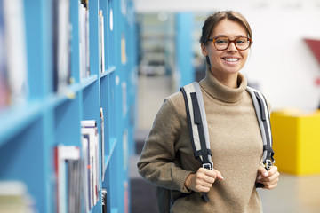Waist up portrait of female college student wearing backpack and smiling at camera cheerfully while posing in library, copy space