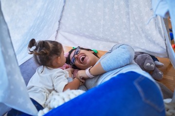 Young beautiful teacher and toddler smiling with happy face lying down over blanket inside tipi at kindergarten