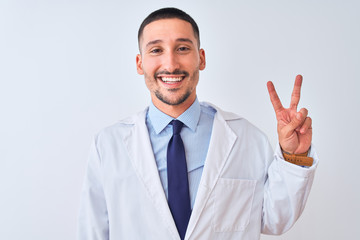 Young doctor man wearing white coat over isolated background smiling with happy face winking at the camera doing victory sign. Number two.