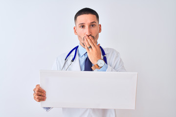Young doctor man holding blank advertising banner over isolated background cover mouth with hand shocked with shame for mistake, expression of fear, scared in silence, secret concept