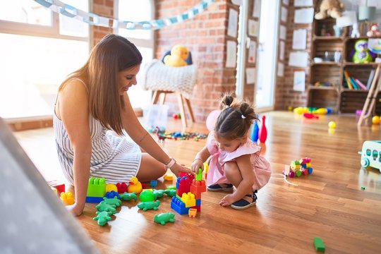 Young beautiful teacher and toddler sitting on the floor playing with building blocks toy at kindergarten
