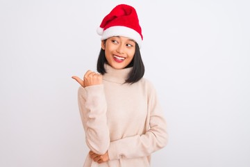 Young beautiful chinese woman wearing Christmas Santa hat over isolated white background smiling with happy face looking and pointing to the side with thumb up.