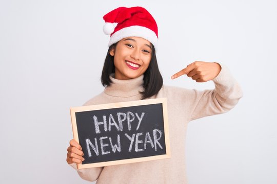 Young chinese woman wearing Santa hat holding blackboard over isolated white background with surprise face pointing finger to himself