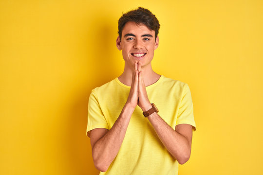 Teenager boy wearing yellow t-shirt over isolated background praying with hands together asking for forgiveness smiling confident.