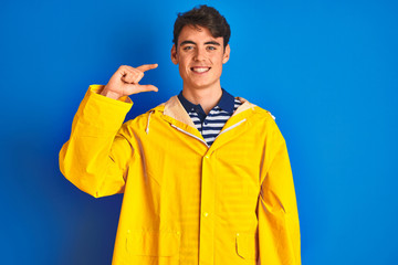 Teenager fisherman boy wearing yellow raincoat over isolated background smiling and confident gesturing with hand doing small size sign with fingers looking and the camera. Measure concept.