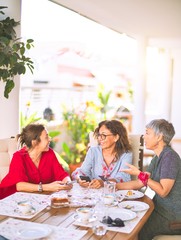 Meeting of middle age women having lunch and drinking coffee. Mature friends smiling happy using smartphone at home on a sunny day