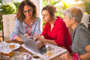 Meeting of middle age women having lunch and drinking coffee. Mature friends smiling happy using laptop at home on a sunny day