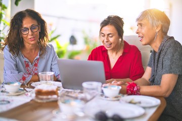 Meeting of middle age women having lunch and drinking coffee. Mature friends smiling happy using laptop at home on a sunny day