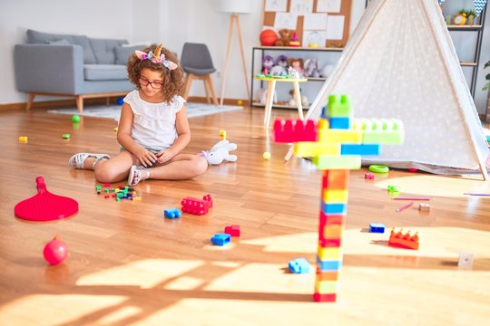 Beautiful toddler wearing glasses and unicorn diadem sitting on the floor playing with building blocks at kindergarten