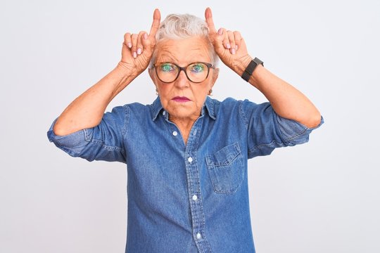 Senior grey-haired woman wearing denim shirt and glasses over isolated white background doing funny gesture with finger over head as bull horns