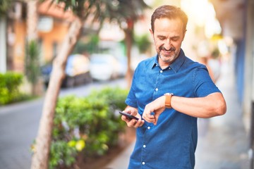 Middle age handsome businessman standing on the street using smartphone looking at the watch