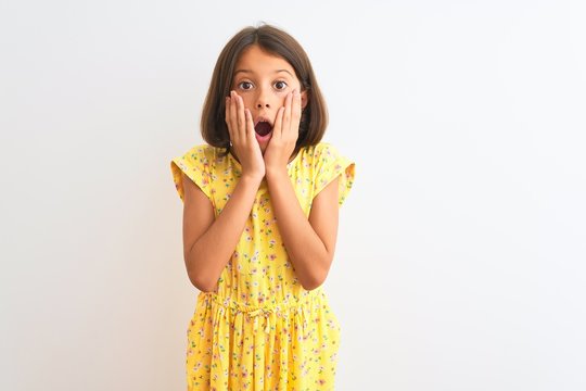 Young beautiful child girl wearing yellow floral dress standing over isolated white background afraid and shocked, surprise and amazed expression with hands on face