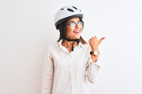 Beautiful businesswoman wearing glasses and bike helmet over isolated white background smiling with happy face looking and pointing to the side with thumb up.