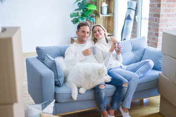 Young beautiful couple with dog sitting on the sofa drinking coffee at new home around cardboard boxes